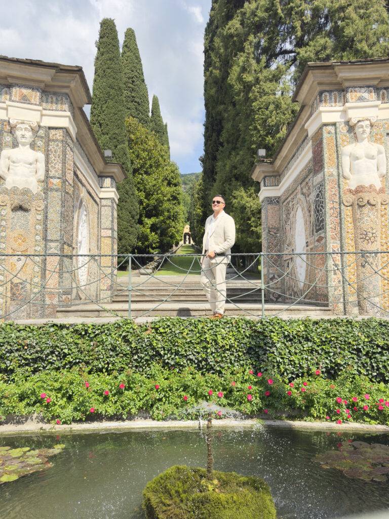 Man in a beige suit posing between mosaic-tiled walls and tall trees in a formal garden.