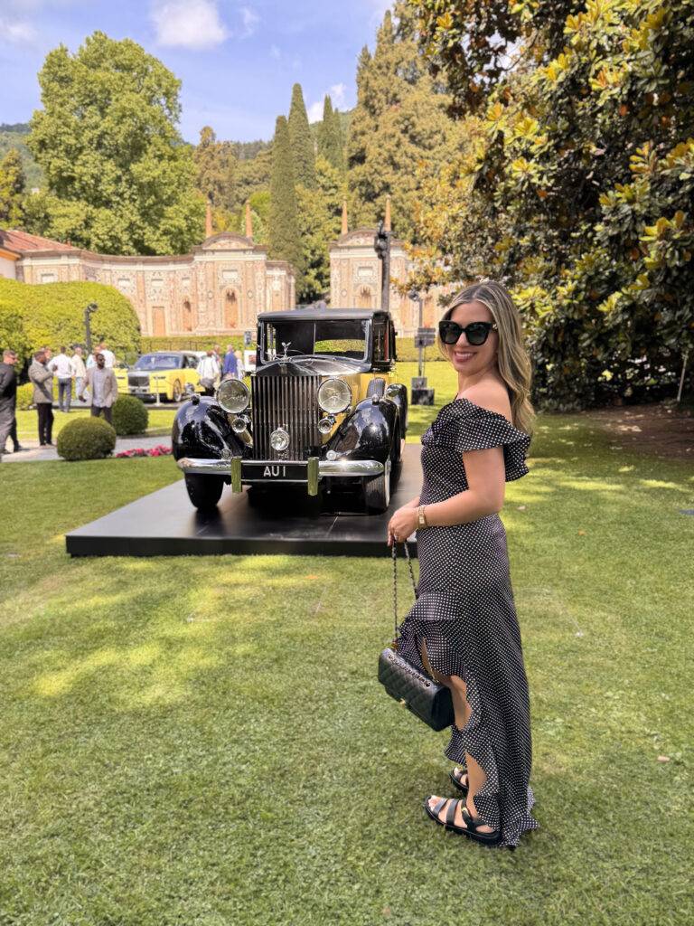 Woman in a black polka-dot dress smiles at camera on a manicured lawn with a vintage black car behind her.