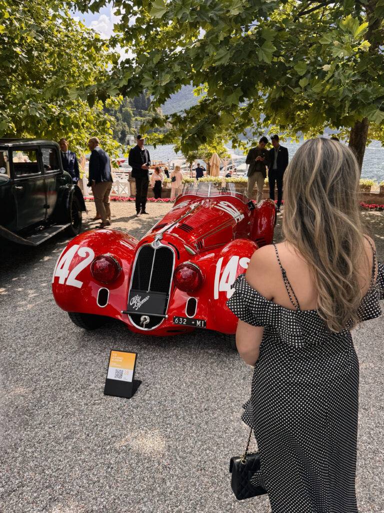 Red vintage race car with number 14 on display near a lakeside promenade, surrounded by spectators under tree shade.