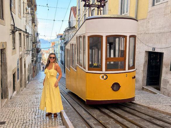 Woman standing beside Lisbon’s iconic yellow elevator tram on a steep cobblestone street, wearing a chic travel outfit that complements the city’s vibrant architecture.
