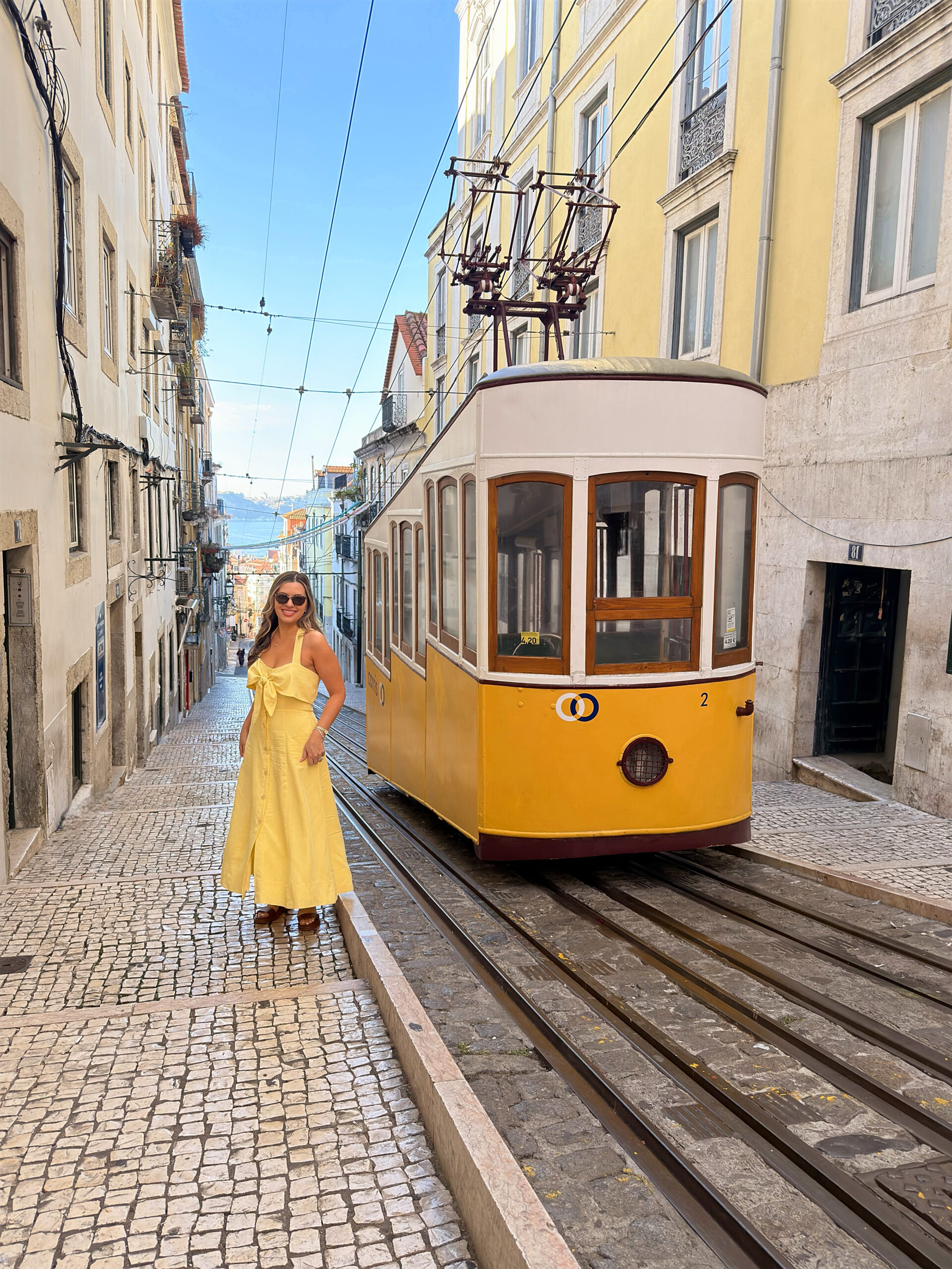 Woman standing beside Lisbon’s iconic yellow elevator tram on a steep cobblestone street, wearing a chic travel outfit that complements the city’s vibrant architecture.
