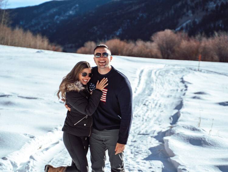 In Aspen wearing chic winter outfits — she’s styled in cozy neutral layers and he’s in a classic Ralph Lauren sweater, surrounded by fresh snow and mountain scenery.