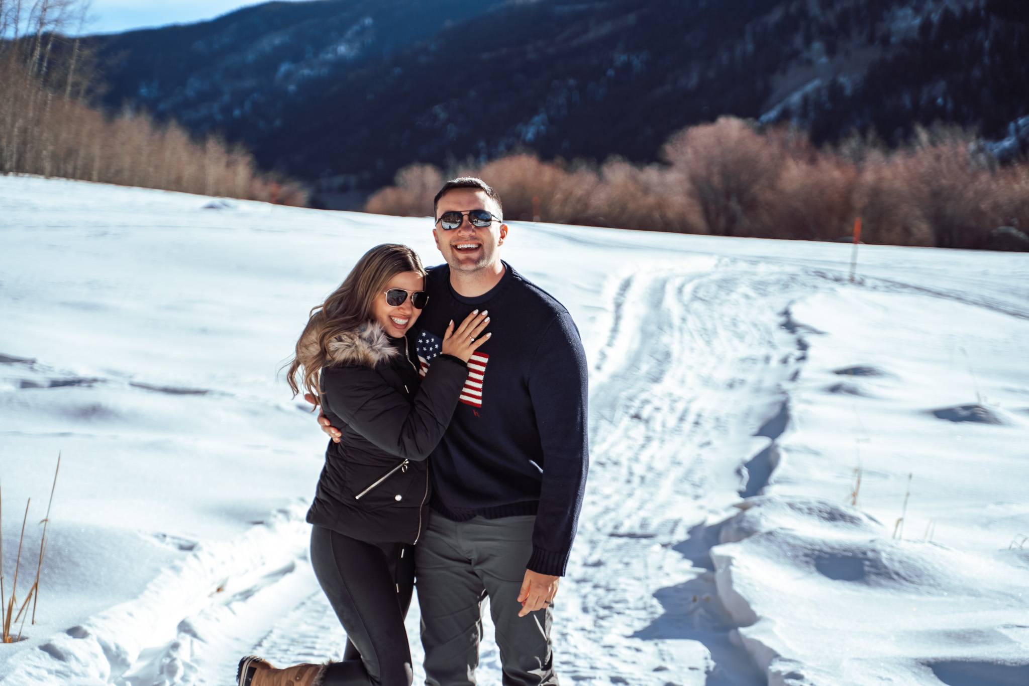 In Aspen wearing chic winter outfits — she’s styled in cozy neutral layers and he’s in a classic Ralph Lauren sweater, surrounded by fresh snow and mountain scenery.