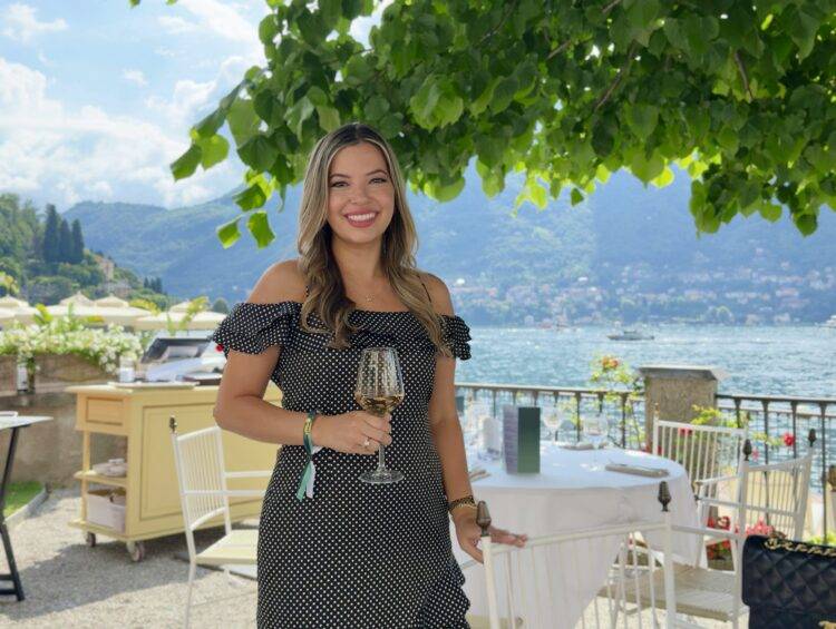 Smiling woman in a black polka-dot dress stands at an outdoor lakeside restaurant on Lake Como with a wine glass, green trees overhead and mountains in the background.