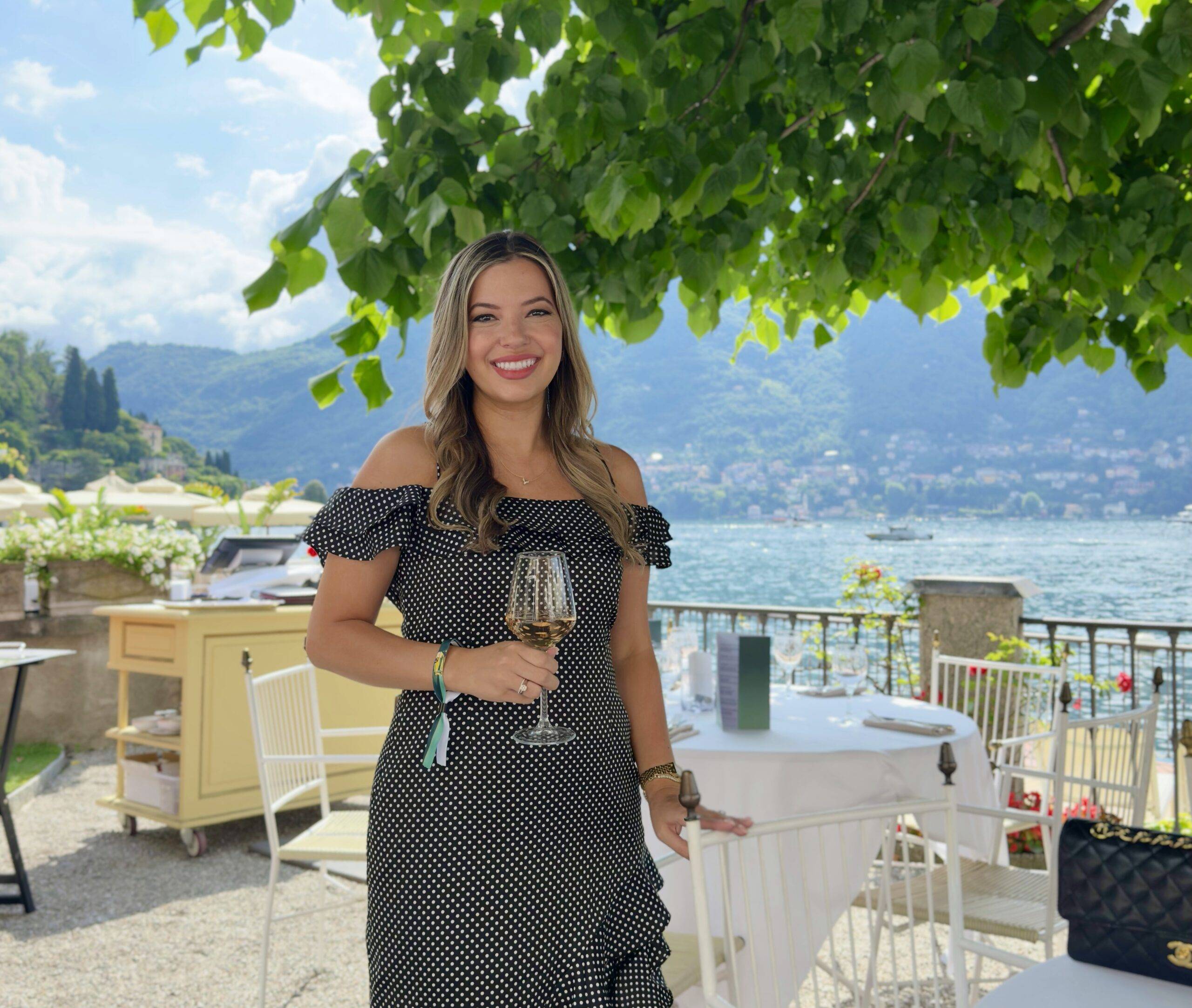 Smiling woman in a black polka-dot dress stands at an outdoor lakeside restaurant on Lake Como with a wine glass, green trees overhead and mountains in the background.